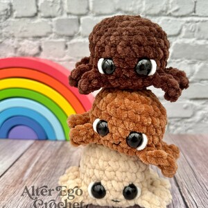 May include: Three crocheted octopus plushies stacked on top of each other. The top octopus is brown, the middle octopus is orange, and the bottom octopus is beige. The octopus plushies have black eyes and a smiling expression. The plushies are in front of a rainbow-colored wooden toy.