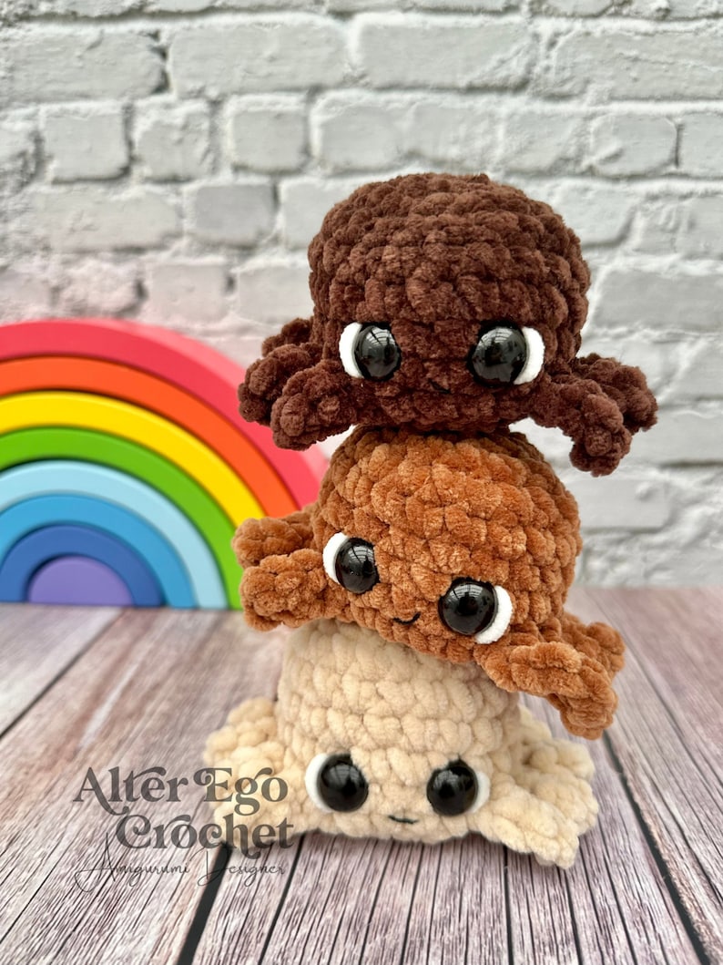 May include: Three crocheted octopus plushies stacked on top of each other. The top octopus is brown, the middle octopus is orange, and the bottom octopus is beige. The octopus plushies have black eyes and a smiling expression. The plushies are in front of a rainbow-colored wooden toy.