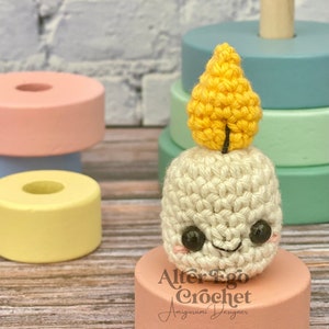 Peut inclure: Une bougie amigurumi crochetée avec un visage souriant, des yeux noirs et des joues roses, surmontée d'une flamme jaune. La bougie repose sur une plateforme rose, avec le texte "Alter Ego Crochet Amigurumi Designer" en dessous.