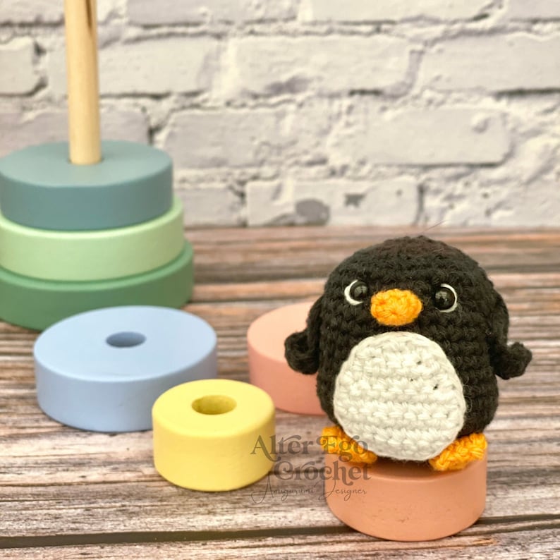 May include: A black and white crocheted penguin toy with an orange beak and eyes. The penguin is sitting on a pink wooden base.