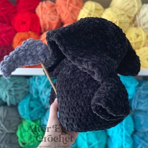 Könnte beinhalten: Ein schwarzes, gehäkeltes Plüschtier in Form einer Kapuzenfigur. Das Spielzeug wird von einer Hand vor einem Regal mit bunten Garnknäueln gehalten. Der Text "After Egg Crochet Amigurumi Designer" ist auf der Hand sichtbar.