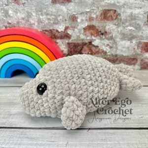 May include: A gray crocheted stuffed manatee with black eyes. The manatee is lying on a wooden surface. The text "Alter Ego Crochet Amigurumi Designer" is visible in the image.