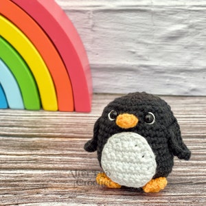 May include: A crocheted penguin toy with black and white fur, an orange beak, and black eyes. The penguin is standing on a wooden surface in front of a rainbow-coloured wooden arch.
