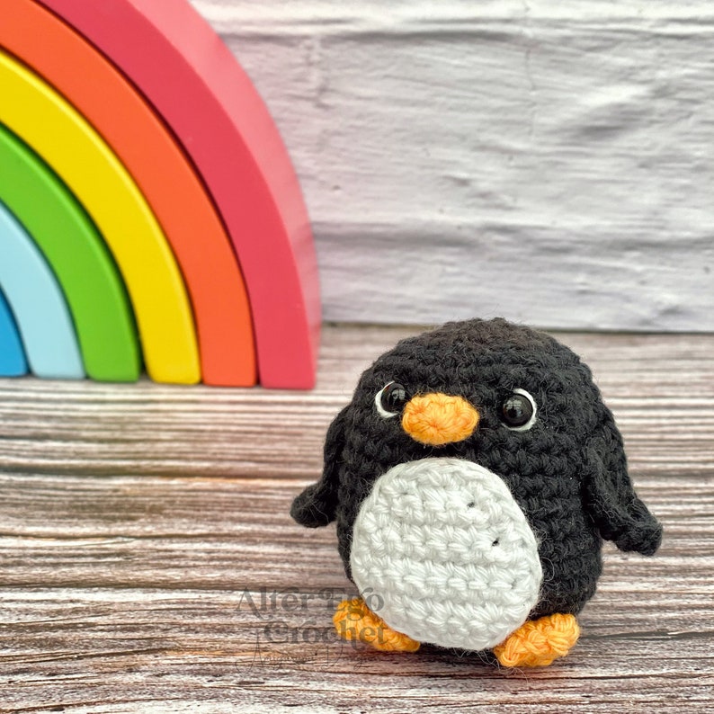 May include: A crocheted penguin toy with black and white fur, an orange beak, and black eyes. The penguin is standing on a wooden surface in front of a rainbow-coloured wooden arch.