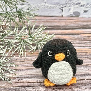 May include: A black and white crocheted penguin toy with an orange beak and feet. The penguin is sitting on a wooden surface with a pine branch in the background.