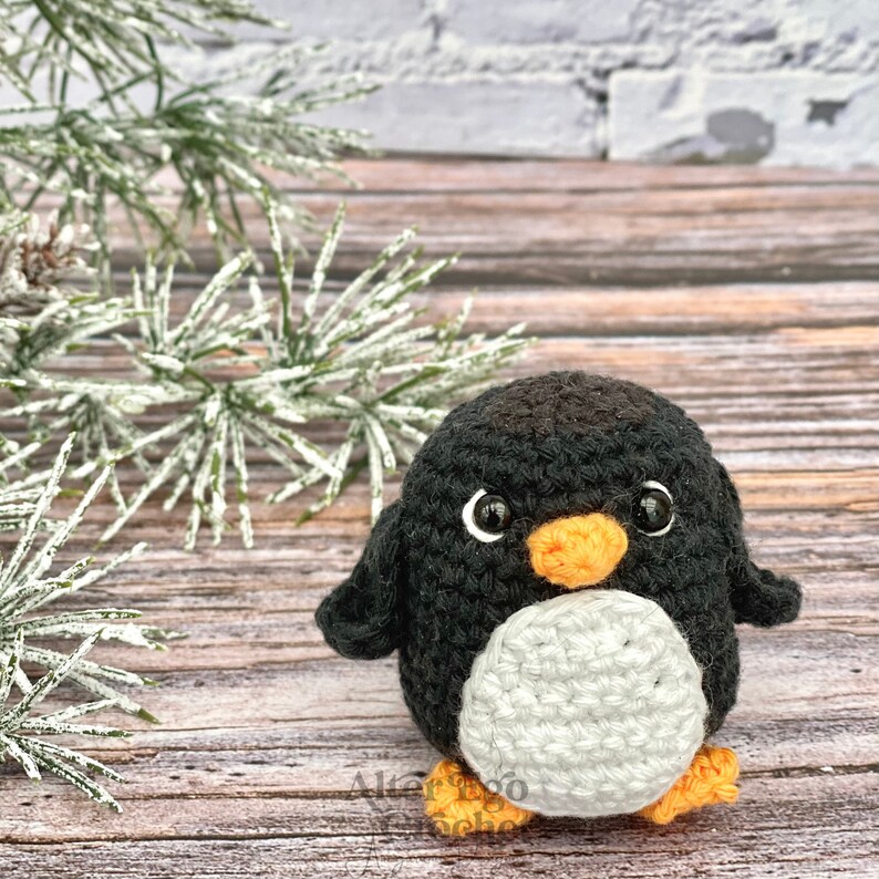 May include: A black and white crocheted penguin toy with an orange beak and feet. The penguin is sitting on a wooden surface with a pine branch in the background.