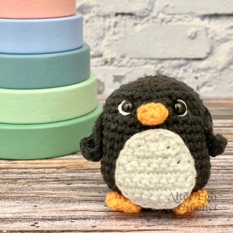 May include: A crocheted penguin toy with black and white fur, an orange beak, and black eyes. The penguin is sitting on a wooden surface.