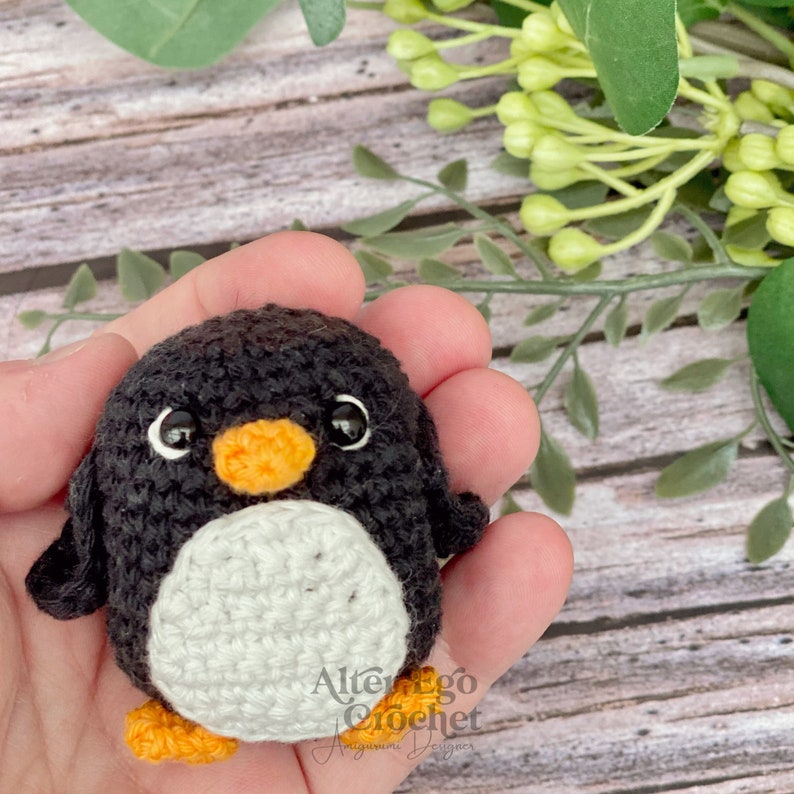 May include: A small crocheted penguin with black and white fur, orange beak and feet. The penguin is being held in a hand.