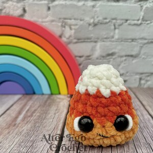 May include: A crocheted candy corn plush toy with a white top, orange middle, and yellow bottom. The toy has black eyes and a small smile. The toy is sitting on a wooden surface in front of a rainbow.