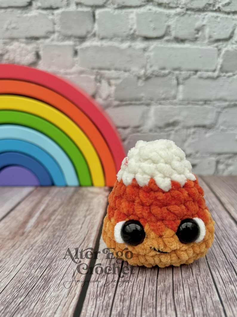 May include: A crocheted candy corn plush toy with a white top, orange middle, and yellow bottom. The toy has black eyes and a small smile. The toy is sitting on a wooden surface in front of a rainbow.