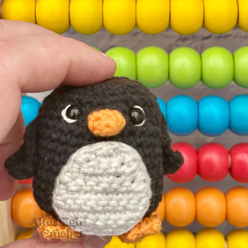 May include: A black and white crocheted penguin with an orange beak and black eyes. The penguin is sitting on a hand in front of a colourful wooden abacus.