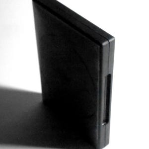 May include: A solid black rectangular object, possibly a case or container, is positioned at an angle against a white background. The object has a smooth, matte finish and a small recessed area on one side.