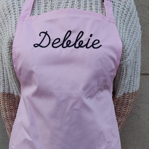 Op de afbeelding: Een roze schort met de naam "Debbie" geborduurd op de voorkant in zwarte cursieve letters.