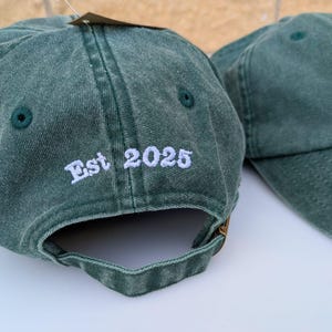 May include: Two dark green baseball caps. One cap has "Est 2025" embroidered in white on the back. The other cap has white text on the front. Both caps have an adjustable strap and a curved brim. The caps are made of a soft, textured fabric.