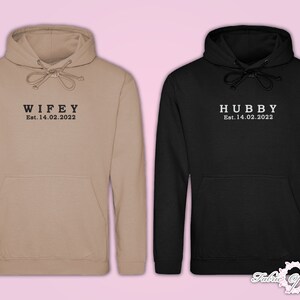 May include: Two hoodies, one beige and one black, with the words "WIFEY Est. 14.02.2022" and "HUBBY Est. 14.02.2022" printed on them.