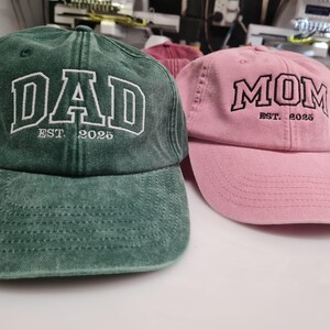 May include: A dark green baseball cap with white embroidered text reading DAD EST. 2025 and a light pink baseball cap with black embroidered text reading MOM EST. 2025. Both caps feature a distressed fabric finish and a curved brim design.