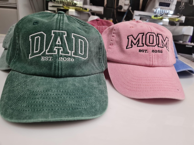 May include: A dark green baseball cap with white embroidered text reading DAD EST. 2025 and a light pink baseball cap with black embroidered text reading MOM EST. 2025. Both caps feature a distressed fabric finish and a curved brim design.