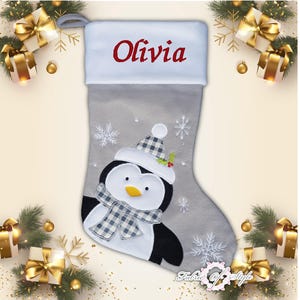 May include: A gray and white Christmas stocking with a penguin design. The stocking has the name "Olivia" embroidered on the cuff.