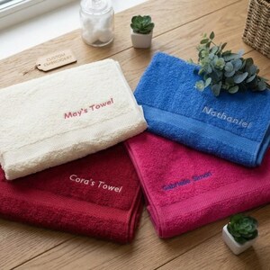 May include: Four folded rectangular towels in cream, dark red, blue, and magenta. Each towel features custom embroidery: cream with 'May's Towel', dark red with 'Cora's Towel', blue with 'Nathaniel', and magenta with 'Gabrielle Simon' in contrasting thread.