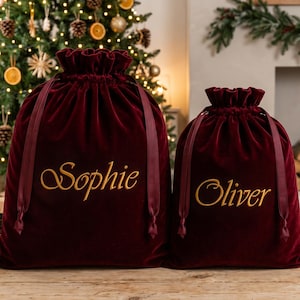May include: Two deep red velvet drawstring gift bags with ruffled tops and matching ribbon closures. The larger bag features the name Sophie embroidered in gold script, while the smaller bag features the name Oliver embroidered in gold script.