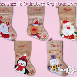 Puede incluir: Cinco calcetines de Navidad personalizados con diferentes diseños bordados. Los calcetines están hechos de arpillera y tienen copos de nieve rojos y blancos. Los nombres "Daddy", "Mummy", "Riley", "Sophie" y "Olivia" están bordados en los calcetines. Los diseños incluyen Papá Noel, un reno, un pingüino y un muñeco de nieve.