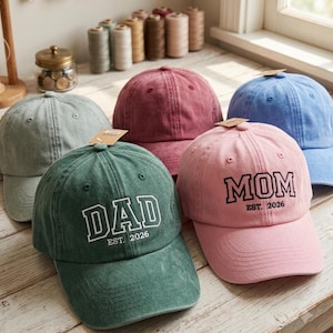 Puede incluir: Una colección de cinco gorras de béisbol en verde salvia, granate, azul, verde oscuro y rosa claro. La gorra verde oscuro tiene texto blanco que dice DAD EST. 2026, y la gorra rosa claro tiene texto negro que dice MOM EST. 2026.