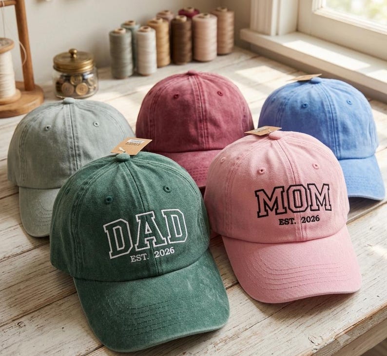 May include: A collection of five baseball caps in sage green, maroon, blue, dark green, and light pink. The dark green cap features white text reading DAD EST. 2026, while the light pink cap features black text reading MOM EST. 2026.
