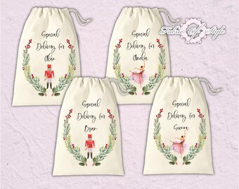May include: Four white cotton drawstring bags with a green wreath design and red berries. Each bag has a different name printed on it: Alan, Amelia, Brian, and Gierra. The bags feature a watercolor illustration of a nutcracker and a ballerina.
