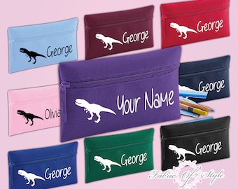 Könnte beinhalten: Neun verschiedene farbige Stiftetuis mit einem Dinosaurier-Motiv und der Möglichkeit, sie mit einem Namen zu personalisieren. Die Farben sind rosa, blau, lila, rot, grün, schwarz, dunkelblau und weinrot. Der Text "9 COLORS AVAILABLE" und "PERSONALISED TO ORDER" ist ebenfalls im Bild enthalten.