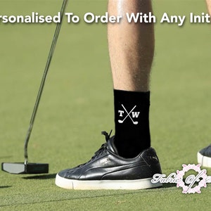 May include: A pair of black socks with white embroidered initials "T" and "W" and crossed golf clubs. The socks are worn by a person on a golf course.