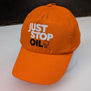 Może przedstawiać: Jasnopomarańczowa czapka bejsbolowa z białym napisem "JUST STOP OIL". Słowo "OIL" znajduje się nad małą grafiką czaszki i skrzyżowanych piszczeli. Czapka ma zakrzywiony daszek i guzik na górze.