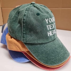 May include: A stack of baseball caps in various colors including green, tan, blue, and red. The top green cap features white embroidered text that reads YOUR TEXT HERE. Each cap has a curved brim, a button on the crown, and stitched eyelets.