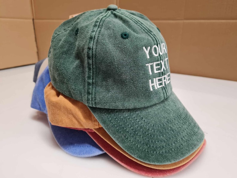 May include: A stack of baseball caps in various colors including green, tan, blue, and red. The top green cap features white embroidered text that reads YOUR TEXT HERE. Each cap has a curved brim, a button on the crown, and stitched eyelets.