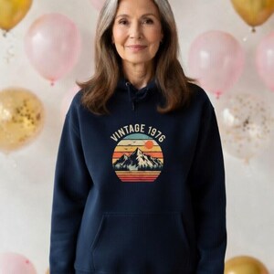 Könnte beinhalten: Ein marineblauer Kapuzenpullover mit einem Vintage-Berg- und Sonnenuntergang-Grafikdesign und dem Text "VINTAGE 1976". Der Kapuzenpullover hat eine Vordertasche und eine Kapuze mit Kordelzug. Die Person auf dem Bild trägt den Kapuzenpullover.
