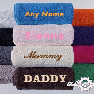 May include: A collection of folded terry cloth towels in various colors including blue, white, black, gray, pink, orange, navy, magenta, green, and brown. Several towels feature embroidered text including Any Name, Sienna, Mummy, and DADDY.