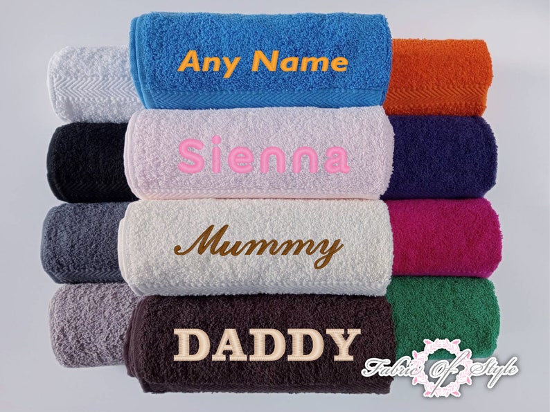May include: A collection of folded terry cloth towels in various colors including blue, white, black, gray, pink, orange, navy, magenta, green, and brown. Several towels feature embroidered text including Any Name, Sienna, Mummy, and DADDY.