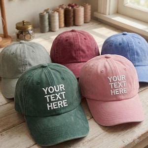 May include: A collection of five baseball caps in various colours including sage green, dusty rose, maroon, light blue, and grey. The green and pink caps feature the white embroidered text "YOUR TEXT HERE" on the front panels.