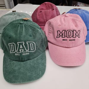 May include: A collection of five baseball caps in various colors including green, pink, blue, and maroon. Each cap features embroidered text on the front, with one green cap reading DAD EST. 2025 and one pink cap reading MOM EST. 2025 in block letters.