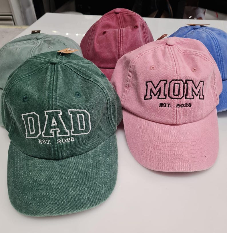 May include: A collection of five baseball caps in various colors including green, pink, blue, and maroon. Each cap features embroidered text on the front, with one green cap reading DAD EST. 2025 and one pink cap reading MOM EST. 2025 in block letters.