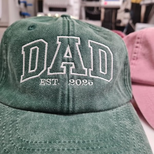 May include: A forest green baseball cap with a distressed, washed-out texture. The front features white embroidered block lettering that reads DAD, with the smaller white embroidered text EST. 2025 positioned directly underneath the main word.