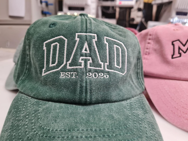 May include: A forest green baseball cap with a distressed, washed-out texture. The front features white embroidered block lettering that reads DAD, with the smaller white embroidered text EST. 2025 positioned directly underneath the main word.