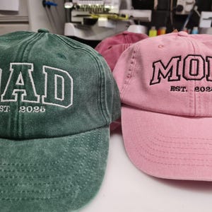 May include: A forest green baseball cap with white embroidered text reading DAD EST. 2025 and a light pink baseball cap with black embroidered text reading MOM EST. 2025. Both caps feature a washed fabric texture and a curved brim design.