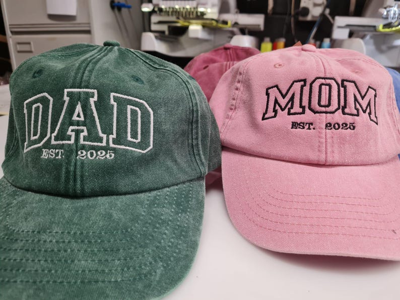 May include: A forest green baseball cap with white embroidered text reading DAD EST. 2025 and a light pink baseball cap with black embroidered text reading MOM EST. 2025. Both caps feature a washed fabric texture and a curved brim design.