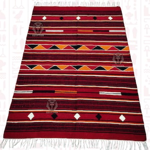 Puede incluir: Alfombra tejida a rayas rojas, negras, blancas y naranjas con un patrón geométrico de diamantes y triángulos. La alfombra tiene flecos en todos los lados.