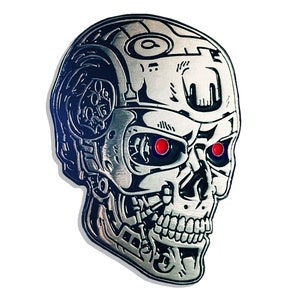 May include: A silver-colored, metallic skull with red eyes, resembling a cyborg. The skull has detailed mechanical features and a shadow, set against a white background. The design is reminiscent of a science fiction theme.