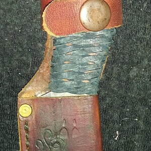 May include: A handmade knife with a dark blue wrapped handle and a brown leather sheath. The sheath features a painted howling wolf design and decorative stitching. A brown leather strap secures the knife.