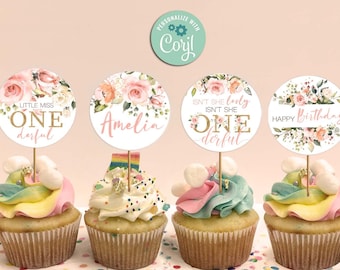 以下が含まれることがあります： 花柄のトッパーが付いた4つのカップケーキ。トッパーには「Little Miss One derful」、「Amelia」、「Isn't She Lovely Isn't She One derful」、「Happy Birthday」と書かれています。カップケーキはカラフルなフロスティングで、紙吹雪で覆われた表面にあります。