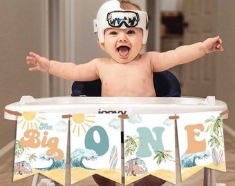 May include: A baby in a walker with a banner that says "The Big One." The banner has a beach theme with a sun, waves, a surfboard, and a vintage van. The baby is wearing a white helmet.