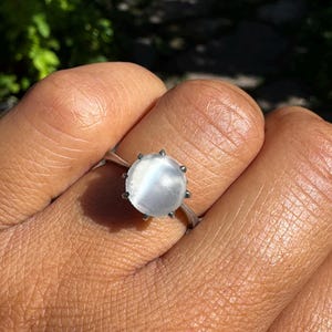 May include: A silver ring featuring a round, translucent white cabochon gemstone with a distinct chatoyant cat's eye effect. The stone is secured in a six-prong setting on a simple, polished silver band that encircles the finger.