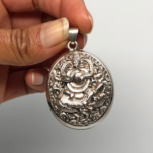May include: Silver pendant featuring an intricate design of a deity surrounded by detailed floral and animal motifs. The pendant is round and has a loop at the top for attaching a chain.